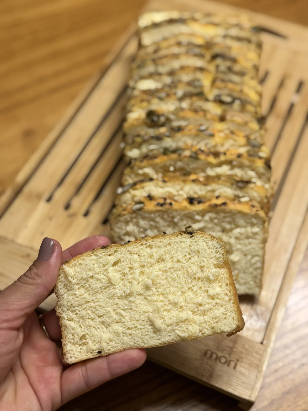 Uma fatia do pão proteico da Victoria nas mãos, com o pão fatiado sobre a tábua